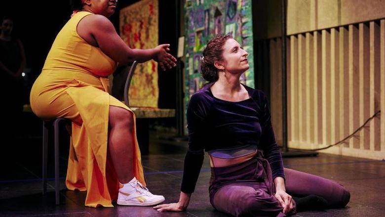 A white woman sits straight on a floor stage next to a Black woman sitting in a chair.